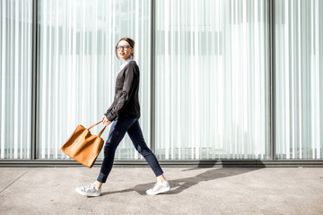 Lifestyle portrait of a young businesswoman walking outdoors with bag on the office wall background