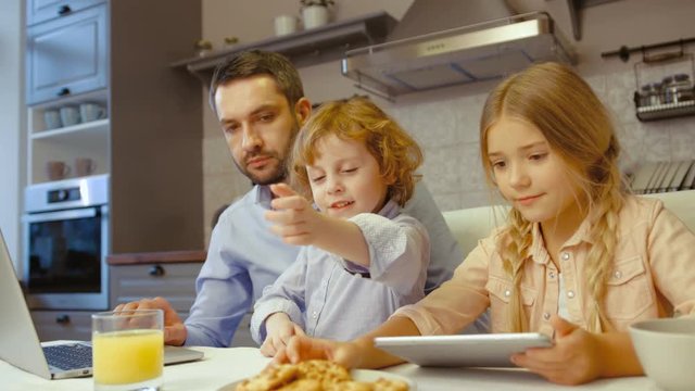 Caucasian man siting in kitchen using laptop and his children siting near him while his wife talking on mobile phone in kitchen.