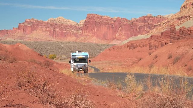 RV camper driving along empty road leading past butte and mesa mountains in red rocky desert on summer day. Trailer home traveling in Capitol Reef Canyon past rocky mountain formations in red desert