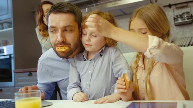 Caucasian handsome man using laptop and his son giving him cookies, his dauther siting children siting beside him with wife and using tablet in kitchen.