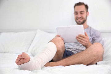 Handsome young man with tablet computer and bandaged leg sitting on bed at home