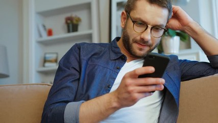 Young caucasian man with glasses and beard relaxing on couch using smart phone in modern industrial house. Indoor. - Powered by Adobe