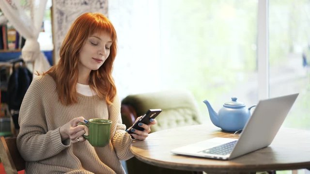 Beautiful red haired woman is sitting in a cafe and drinking tea from a large green mug. Laptop in front of her on the table. She is web surfing with her smartphone. Locked down real time medium shot.
