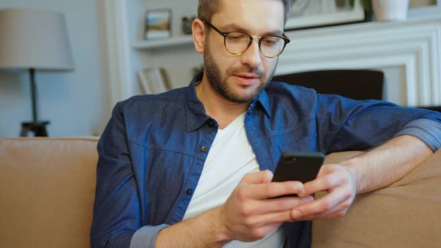 Attractive caucasian man with glasses and beard siting on couch using mobile phone for playing game in living room. Indoor.