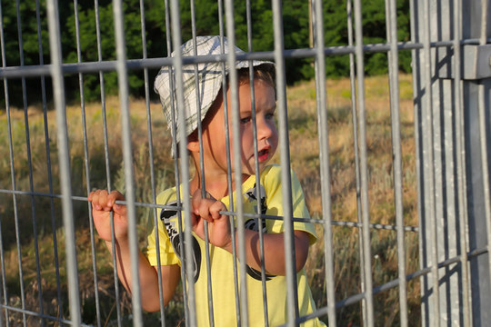 A little white boy in a T-shirt with painted headphones behind an iron grating fence. The kid behind the lattice fence looks anxiously aside.