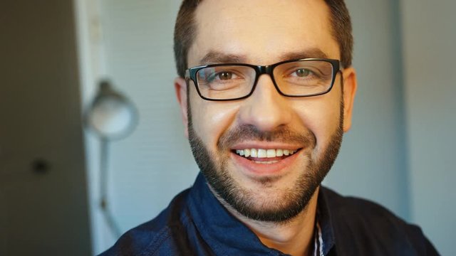 Portrait of young caucasian beautiful man in glasses with beard posing to the camera and smiling with teeth. Indoor.