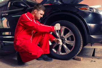 Young concentrated male auto mechanic sitting near a black sedan and screwing car wheel