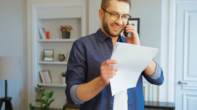 Handsome caucasian business man in glasses in stylish modern office celling on smart phone and holding in hands sheets of paper. Indoor..