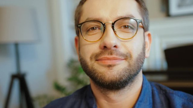 Portrait of caucasian attractive man in glasses with beard in blue shirt looking to the camera and smiling. Indoor.