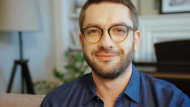 Portrait of caucasian young man in glasses with beard in blue shirt looking in side, turning to the camera and smiling. Indoor.