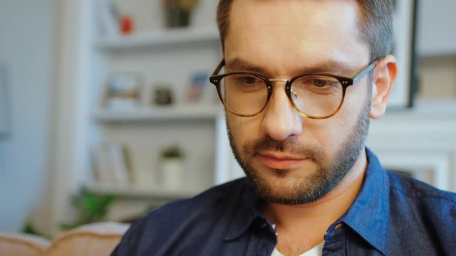 Portrait of concentrated caucasian attractive man in glasses with beard and dark hair in blue shirt. Indoor.