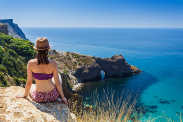 Beautiful woman sitting on a cliff above Black sea and looking at deep blue water. Feel free to explore, adventure and discovery nature treasure.From the back. Top view. Tourism and travell.