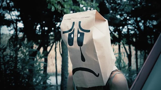 A breadbag face at a public park, sitting on a slide, with a sad depressed expression. Close-up shot.

