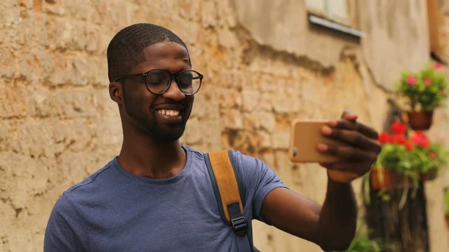 Handsome young african american man in glasses video chatting with friend using mobile phone on city background.
