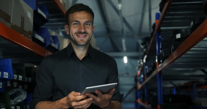 Young attractive delivery logistics male in strict clothes standing in a hardware warehouse with a tablet in his hand checking information. Camera back movement, lights switching off, man smiling