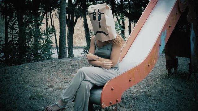 A breadbag face at a public park, sitting on a slide, with a sad depressed expression. Medium shot.
