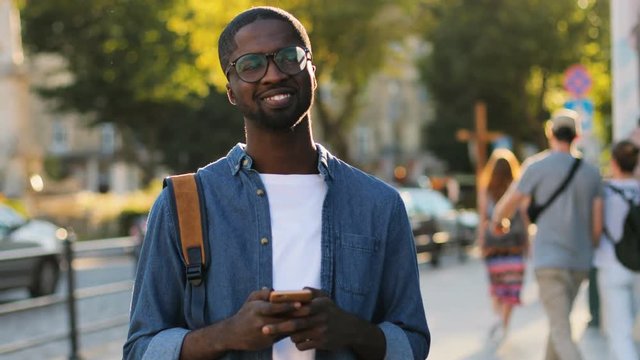 Young african tourist in blue shirt using smart phone, looking to the camera and smiling with teeth standing city center. Outdoor.