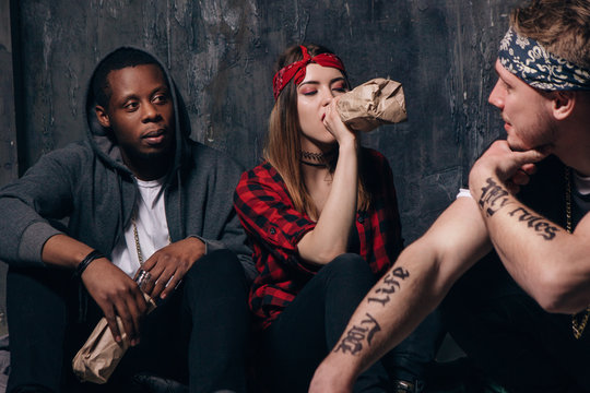Group of alcoholic young people on night party. Youth addiction problem. Tired drunk caucasian girl drink alcohol from bottle, sad guys with tattoo sits near . Students lifestyle