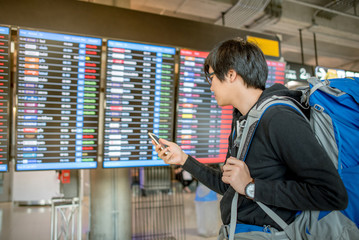 Young asian man with backpack bag checking for his flight from the flight information board and holding smartphone at airport terminal