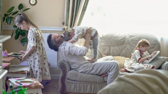 Wide shot of senior man with grey bushy beard dressed in traditional Russian clothing sitting on sofa and lifting laughing little boy in air; little girls in simple dresses playing on tablets