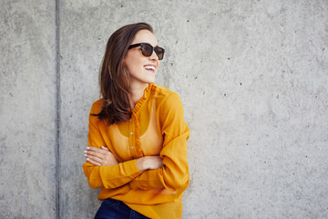 Beautiful happy lady leaning against concrete wall and looking away in sunglasses