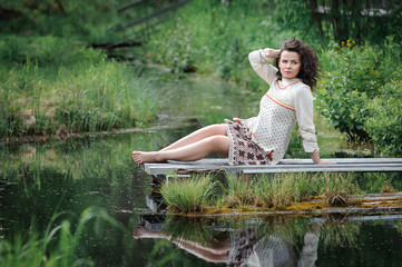 Portrait of a beautiful girl in Slavic clothes. Young barefoot woman is sitting by the pond. Summertime