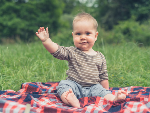 Cute little baby in nature on picnic blanket is waving