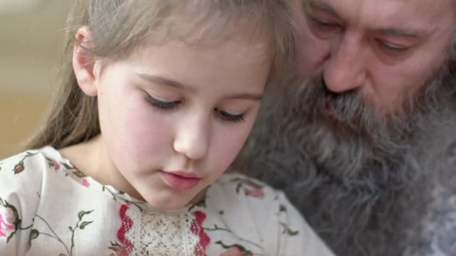 Close up of little girl doing her homework with concentration while senior man with grey beard explaining her something