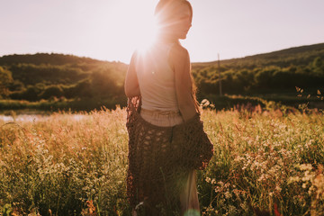 Young brunette woman standing in the sunlit evening field wearing bo ho clothes. Attractive girl in sunset. Girl walking in meadow in evening. Outdoor