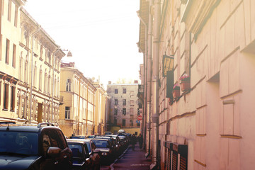 Obraz premium City street at dawn, illuminated by the sun, rows of houses and cars, random passers-by