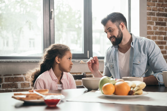 portrait of father feeding grumpy daughter with porridge during breakfast