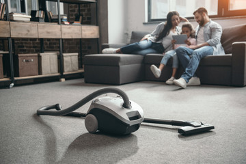 family sitting on sofa and using digital tablet, selective focus on vacuum cleaner