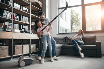 father with daughter holding vacuum cleaner while mother sitting at sofa at home