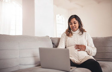 Beautiful pregnant woman with laptop sitting on sofa