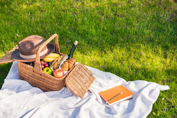 Close-up view of picnic basket with fruits and wine, hat and notebook with pencil on grass