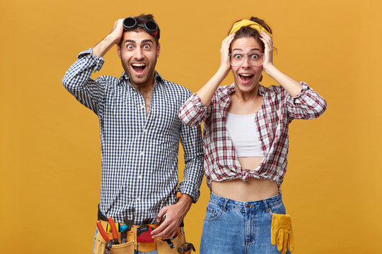 Two workers of maintenance service posing at studio wall, looking at camera in excitement, touching heads and screaming with mouths wide opened. Repairing, remodeling and renovation concept