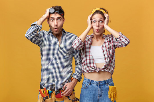 Human emotions and feelings. Two surprised astonished young Caucasian service technicians wearing safety eyeglasses and overalls having amazed and shocked looks, holding hands on their heads