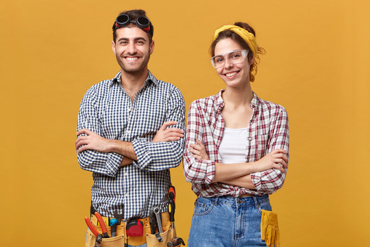 People, lifestyle, job and occupation concept. Studio portrait of happy confident female electrical technician in safety goggles posing in studio together with her male colleague, both folding arms