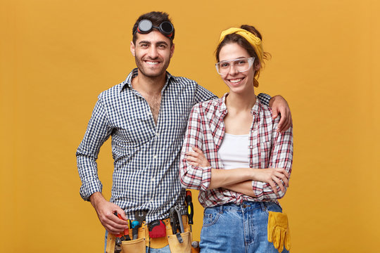 Isolated shot of attractive confident young European employees of maintenance dressed in overalls and protective wear, equipped with instruments, ready for work, looking at camera with happy smiles