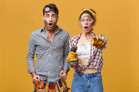 Surprised couple of male and female electical technicians in protective goggles and overalls having astonished looks, girl with drill pointing index finger at camera, showing something shocking