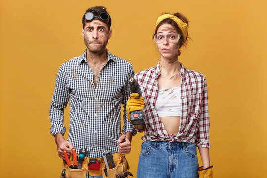 Human facial expressions and emotions. Indoor shot of astonished young male electrician wearing belt knit with instruments posing in studio with confused woman with drill, both looking puzzled