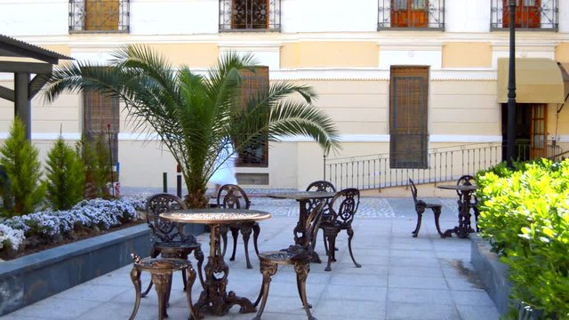 This is footage of a palza with iron chairs and iron tables set to to be used for this small plaza in a spanish traditional fashion. Some people walk by.