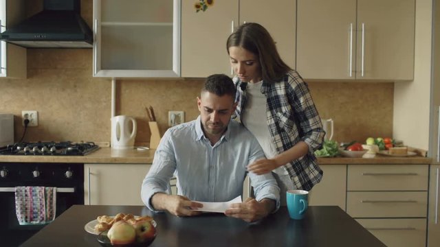 Uspet young man reading unpaid bills and hugged by his wife supporting him in the kitchen at home