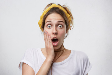 Headshot of young attractive woman with bugged eyes and widely opened mouth holding her hand on...