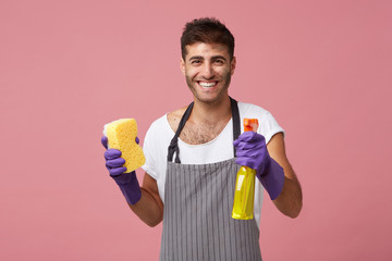 Waist-up portrait of happy positive young man with beard smiling broadly while doing housework by...