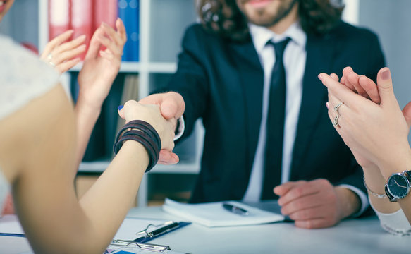 Close up businessmen handshake on team meeting with clapping group of people in background at modern startup business office interior.
