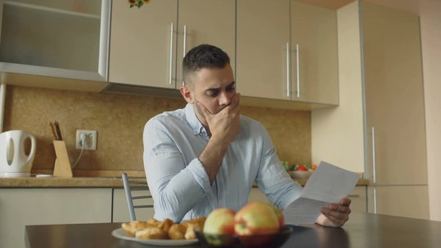 Uspet young man reading unpaid bills and hugged by his wife supporting him in the kitchen at home