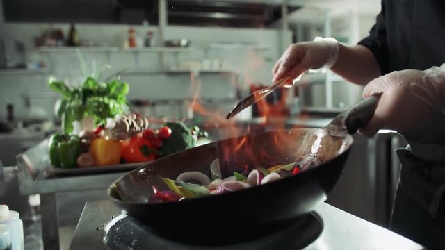 close-up of a chef working the wok with flames roasting mixed colorful vegetables tossing them , restaurant kitchen , slow motion