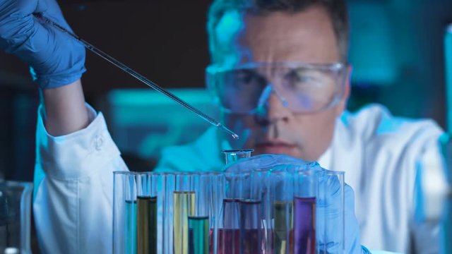 Man scientist or technologist doing experiment in a laboratory pipetting a chemical solution from a beaker held in him gloved hand as he sits behind a row of test tubes in a rack.