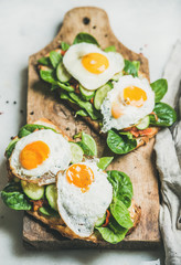 Healthy breakfast sandwiches. Bread toasts with fried eggs and fresh vegetables on wooden board over grey marble background, selective focus. Clean eating, healthy, weight loss, detox food concept